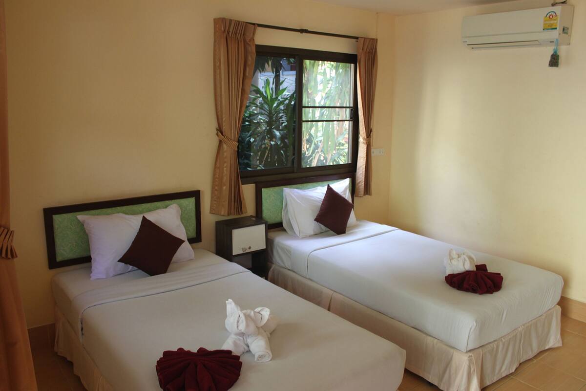 Dos camas individuales con sábanas blancas y almohadas marrones, una ventana con cortinas y un equipo de aire acondicionado en la pared.