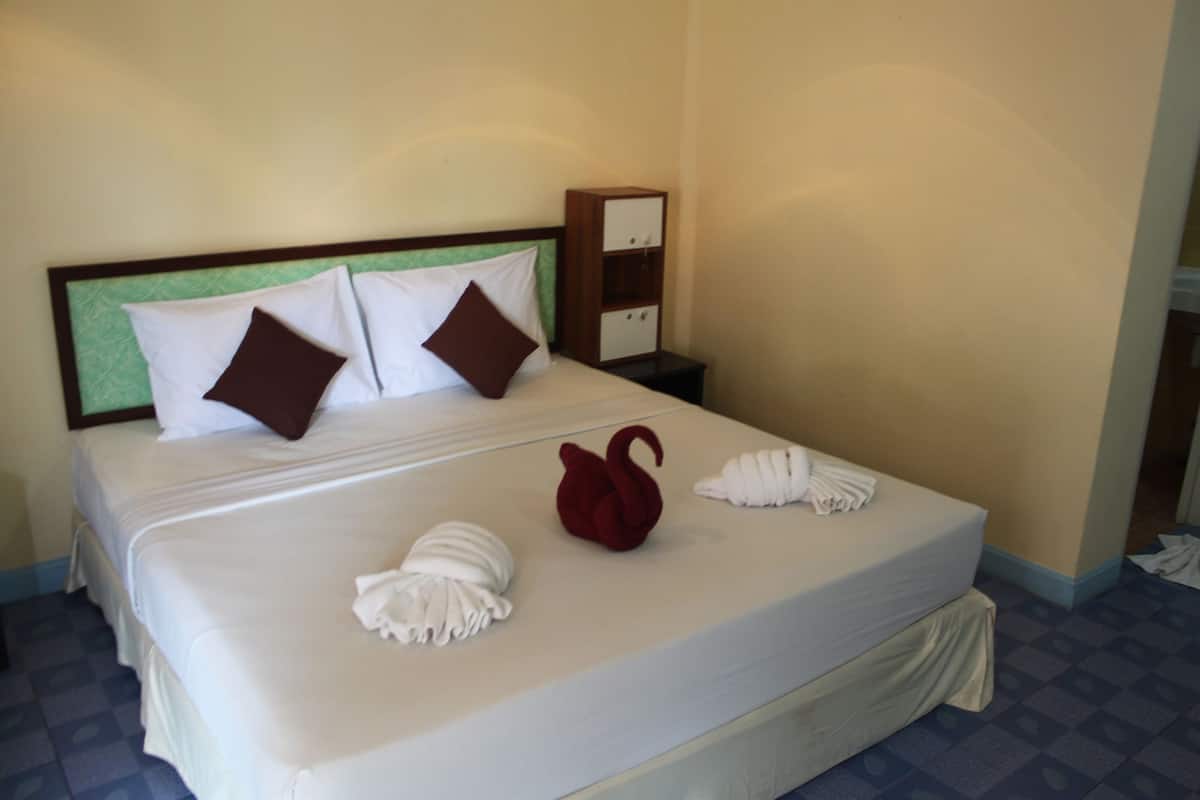Una cama bien hecha con almohadas blancas y marrones, una toalla con forma de cisne rojo y una toalla con forma de cisne blanca. Hay una mesita de noche de madera con un espejo y un estante.