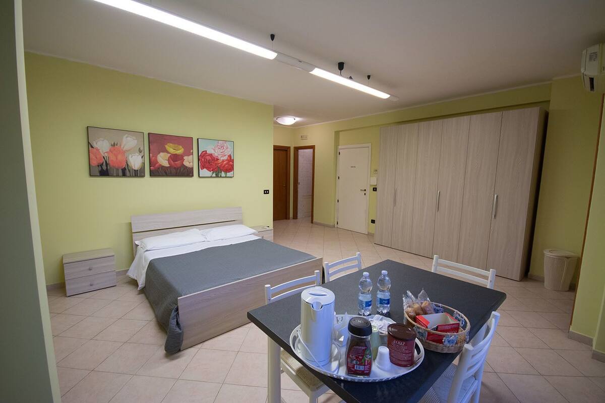 A bedroom with a bed, bedside tables, and a small desk with a chair. There are three paintings on the wall, a wardrobe, and a trash bin.
