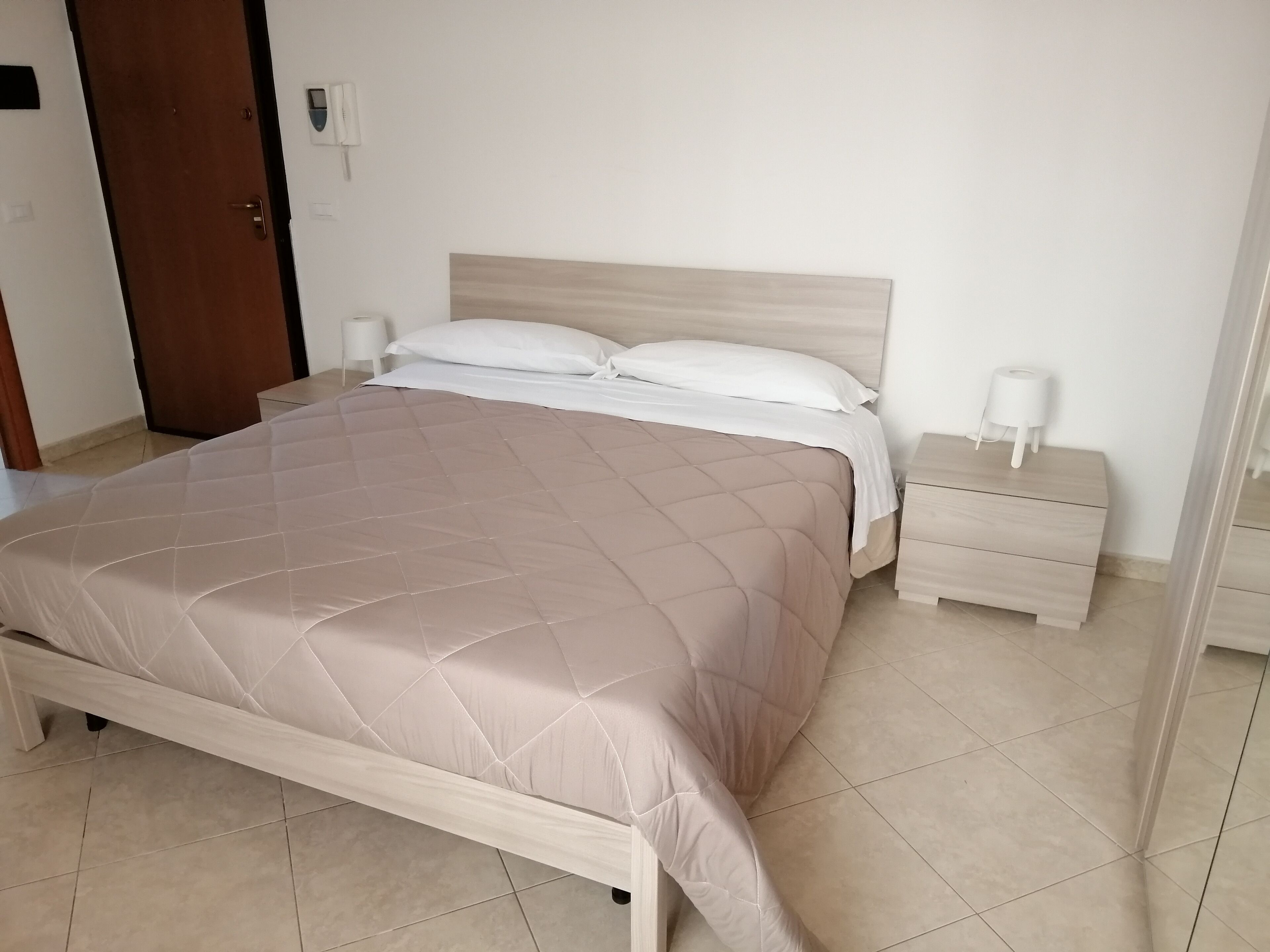 Een net opgemaakt bed met een beige deken, een houten hoofdbord en twee nachtkastjes met lampen.