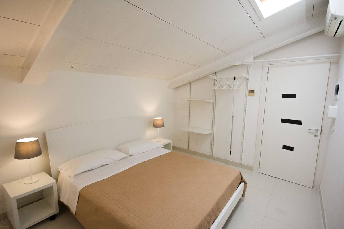 A bedroom with a bed, two bedside tables with lamps, a wardrobe, and a window.