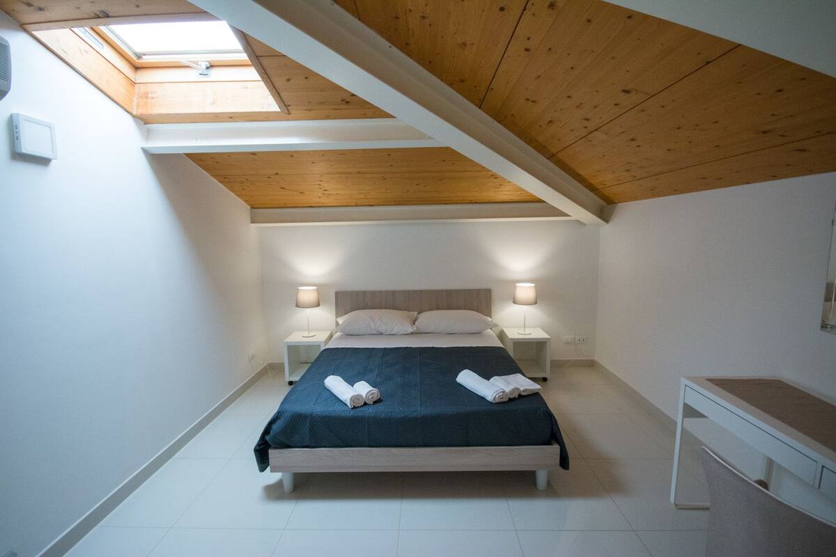 A bedroom with a skylight, a bed with two pillows, two bedside lamps, and a bedside table with towels.