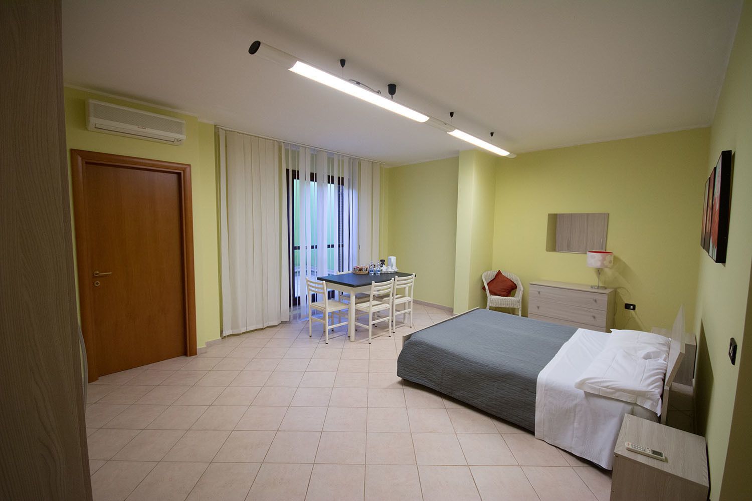 Habitación de hotel con cama, escritorio y silla. Se ve una ventana con cortinas, una puerta y una lámpara de pared.