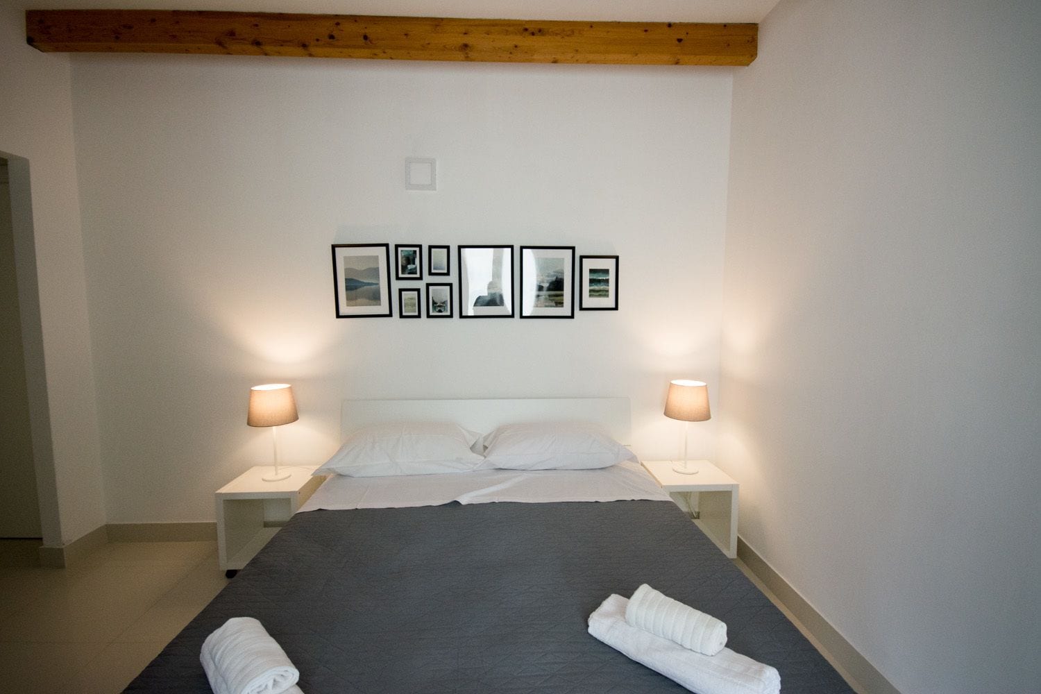 Una cama bien hecha con sábanas blancas, dos lámparas de noche y una serie de cuadros enmarcados en la pared.