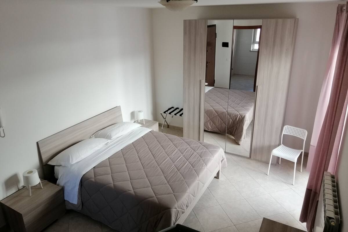 A modern hotel room with a large bed, a bedside table with a lamp, a chair, and a mirror reflecting the room.