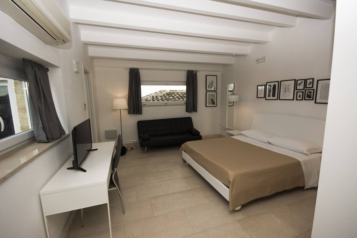 A hotel room with a bed, a desk, a chair, a TV, a window with curtains, and framed pictures on the wall.