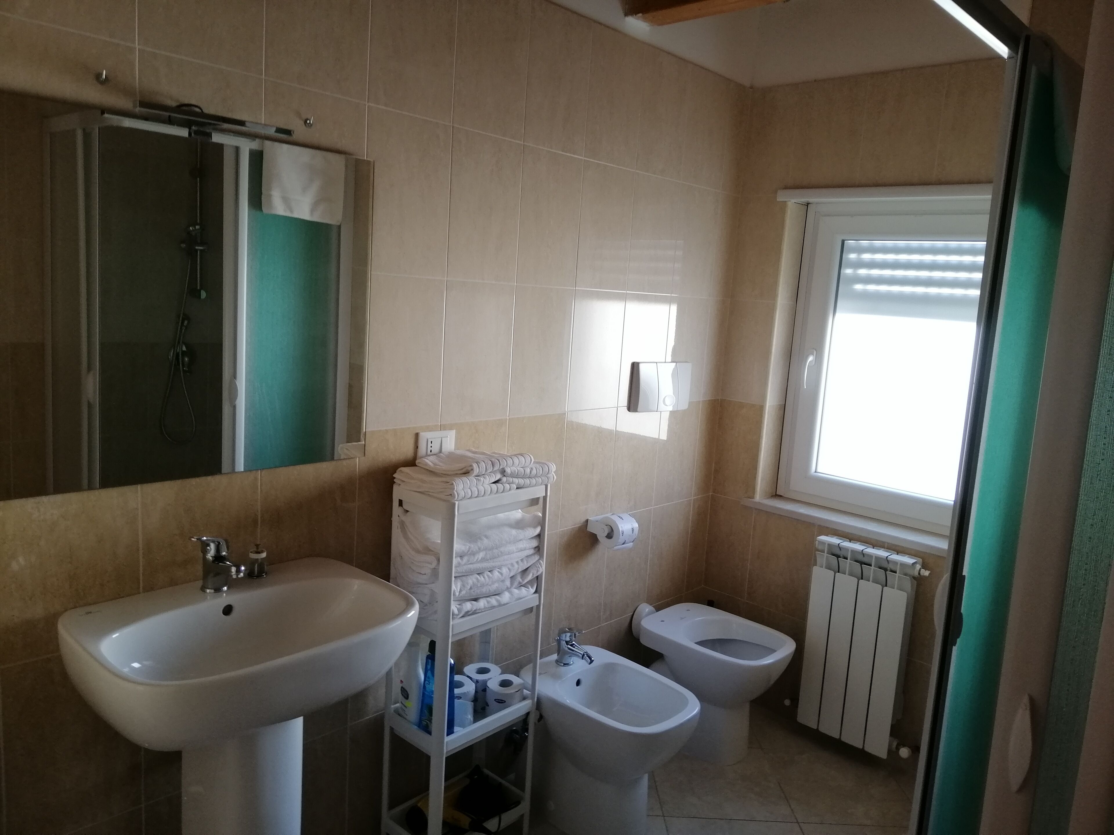 Un baño con lavamanos, bidé y espejo. Se ve un perchero con toallas y un estante con artículos de aseo.