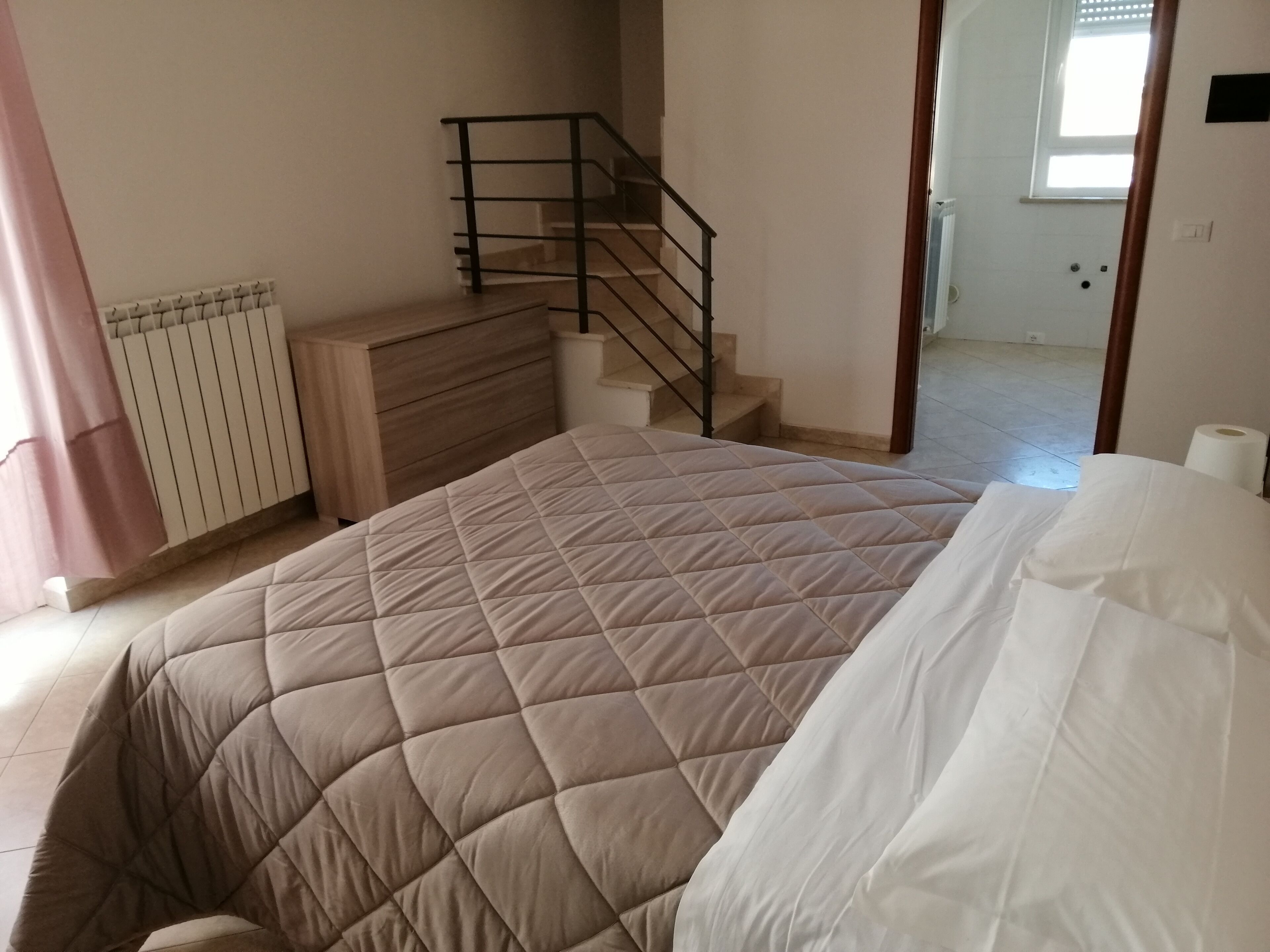A bedroom with a bed, a dresser, a radiator, a staircase, and a window.