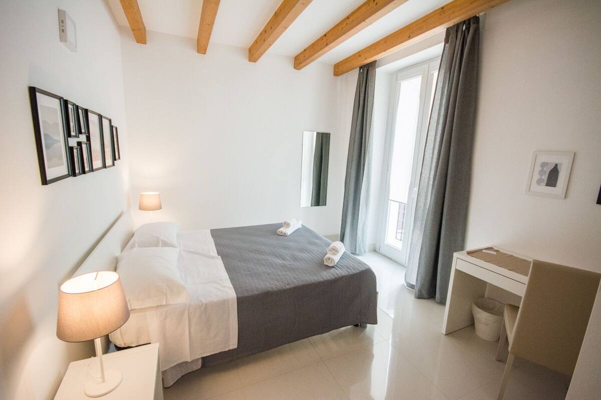 A modern hotel room with a bed, bedside table, lamp, and a desk with a chair.