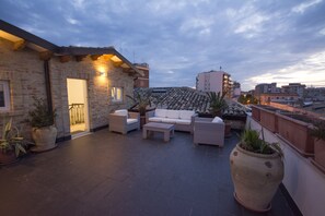 Una terraza en la azotea con mobiliario exterior, plantas en macetas y un edificio de ladrillo.