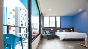 Una habitación de hotel moderna con una cama grande, un pequeño sofá, una mesa de comedor y un balcón con sillas azules.