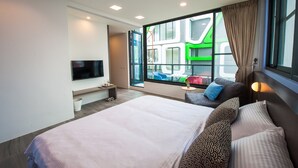 Habitación de hotel moderna con una cama grande, un televisor montado en la pared, una mesita pequeña y una ventana con vistas a edificios coloridos.