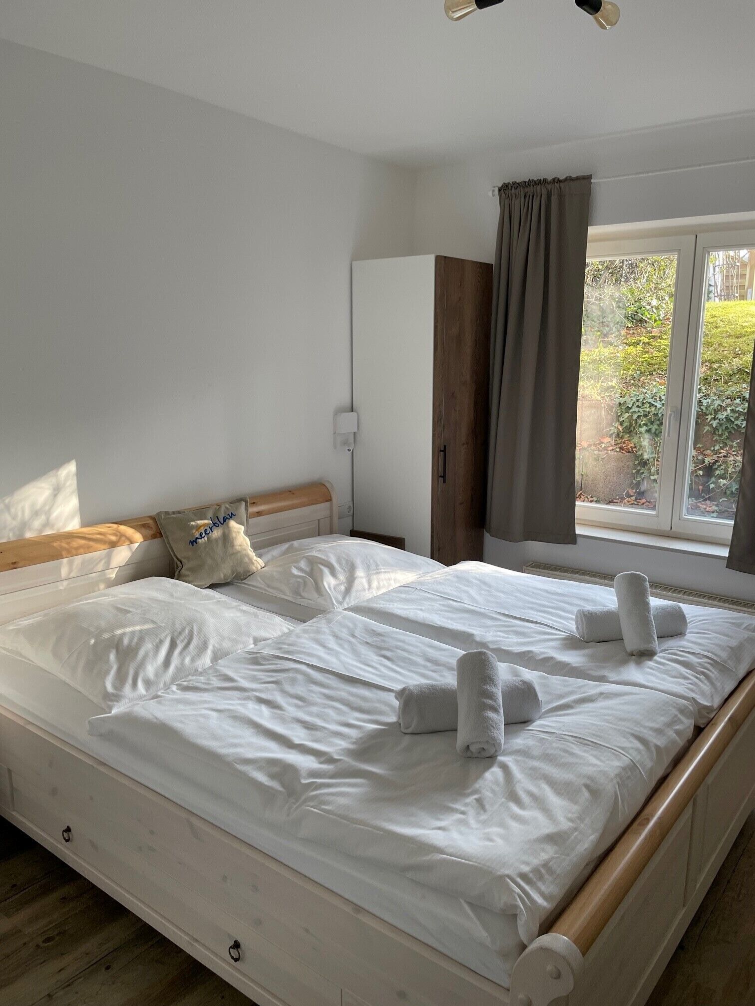 A neatly made bed with white linens and rolled towels, a wooden headboard, a window with curtains, and a white wardrobe.