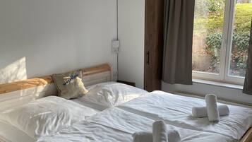 A neatly made bed with white linens and rolled towels, a wooden headboard, a window with curtains, and a white wardrobe.
