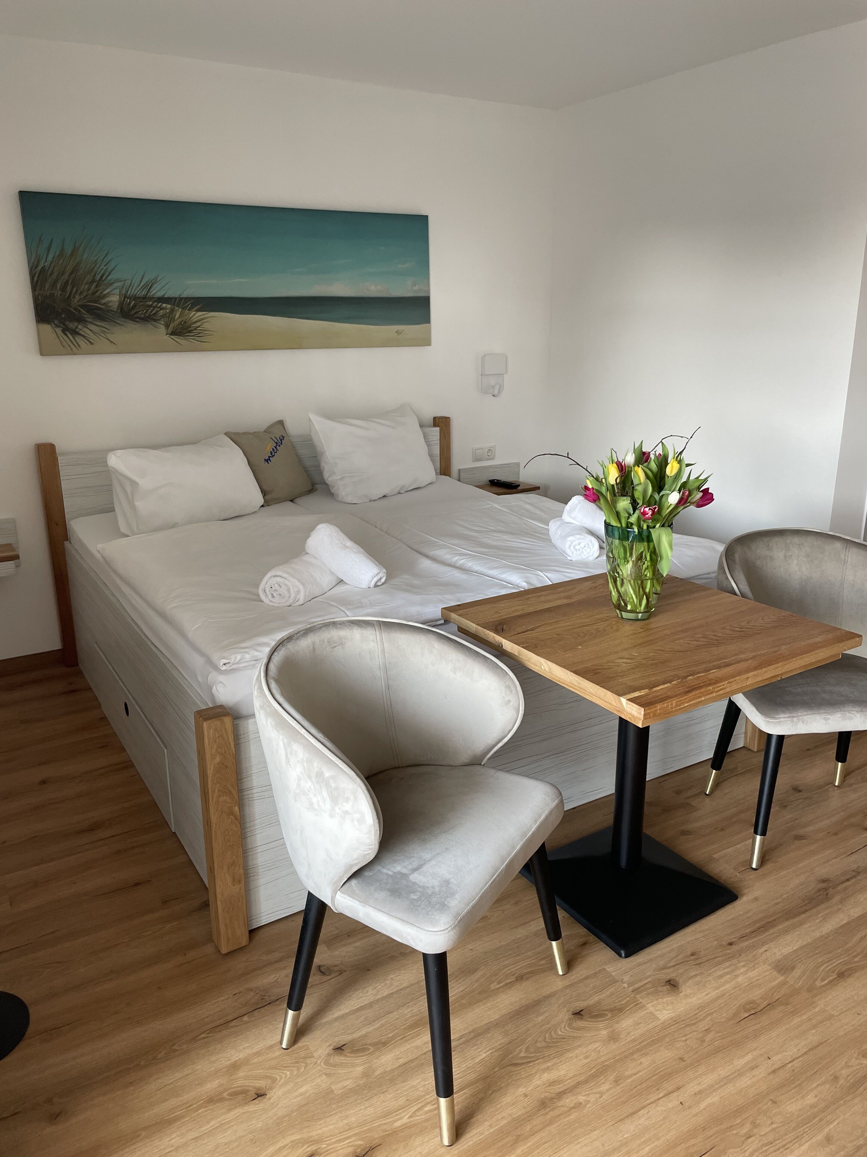 A bedroom with a bed, a wooden table, two chairs, a vase with flowers, and a painting on the wall.