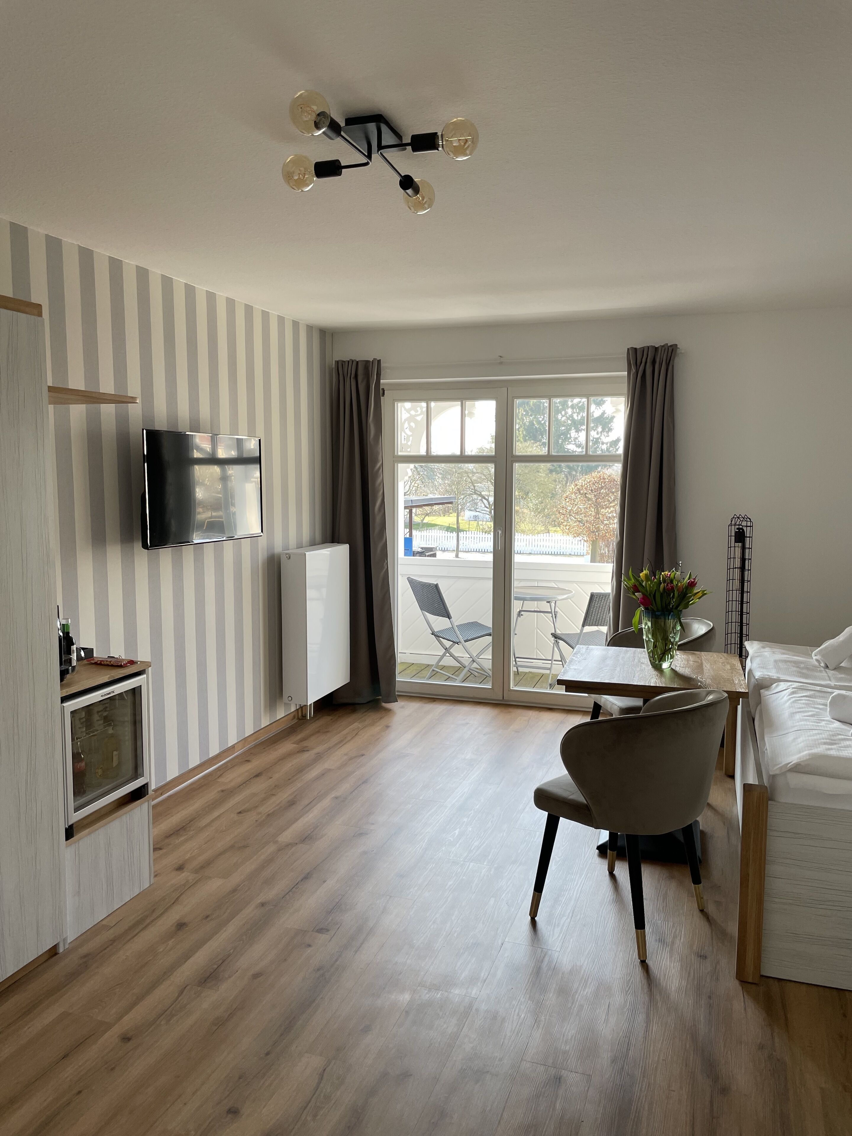 Ein modernes Hotelzimmer mit Flachbildfernseher, einer kleinen Küchenzeile, einem Essbereich mit Tisch und Stühlen und einer großen Schiebeglasdoor, die auf eine Außenpatio führt.