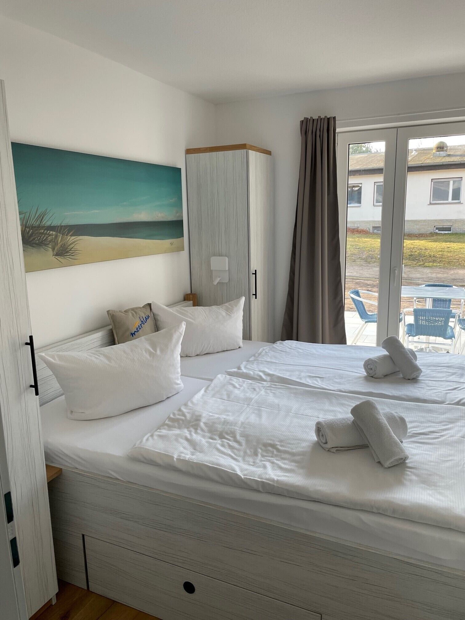 A bedroom with a bed, pillows, a painting, a wardrobe, and a view of the outside.