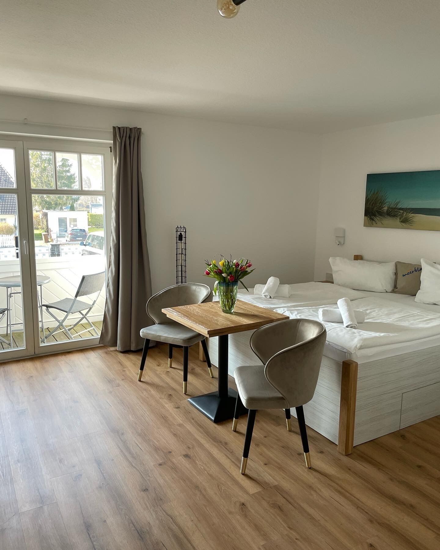 Quarto de hotel moderno com piso de madeira, uma mesa de jantar com cadeiras, uma cama com roupa de cama branca e um quadro na parede.