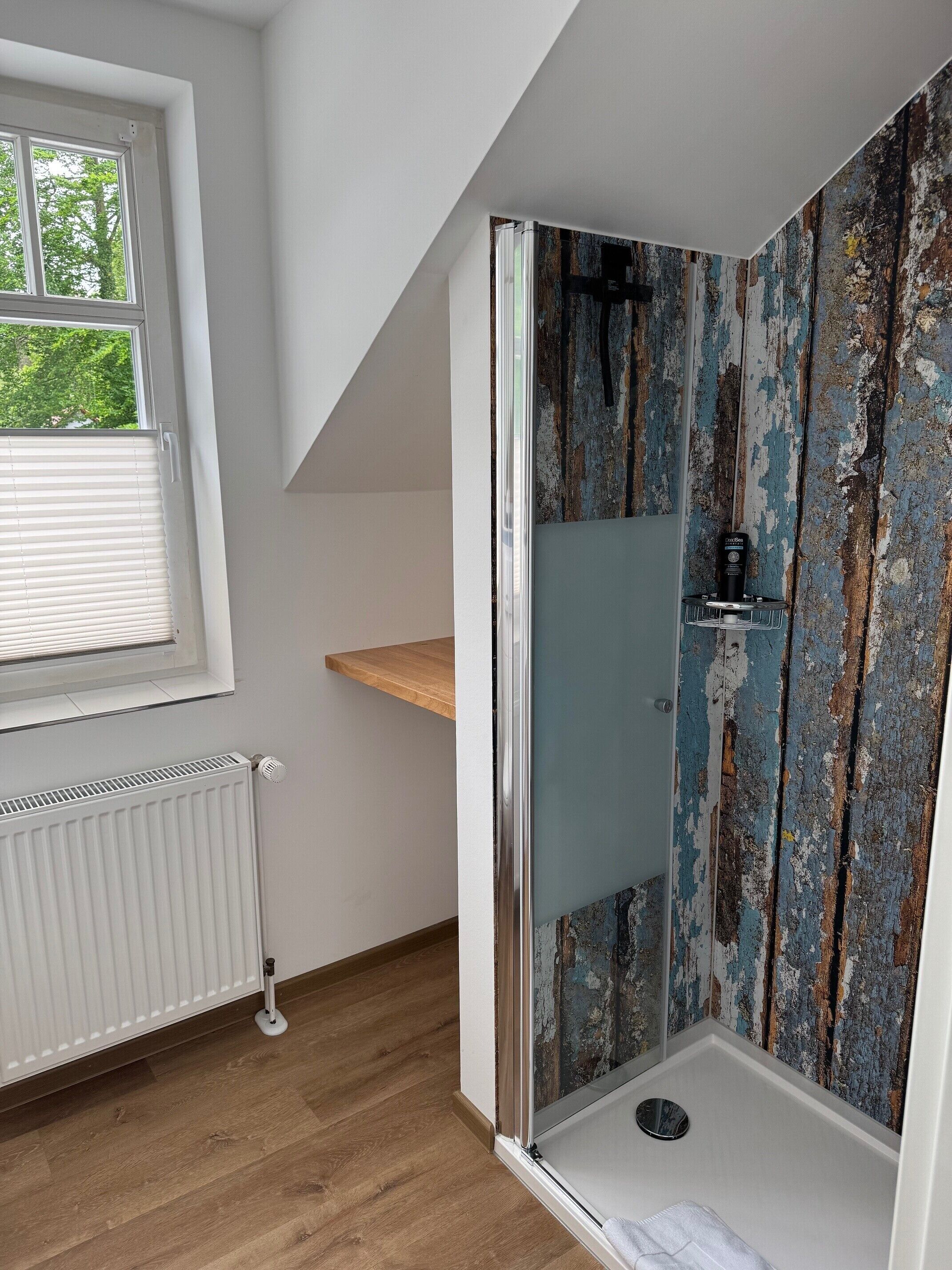 A bathroom with a shower, a window, a radiator, and a wooden shelf.