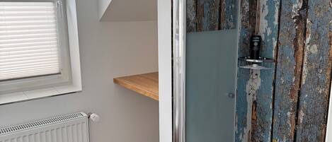 A bathroom with a shower, a window, a radiator, and a wooden shelf.