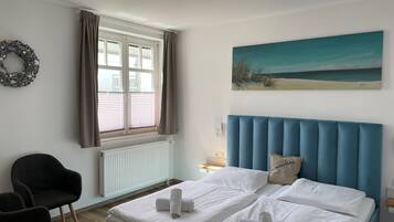 Un dormitorio con cama, una silla, ventana con cortinas, un radiador y un cuadro en la pared.