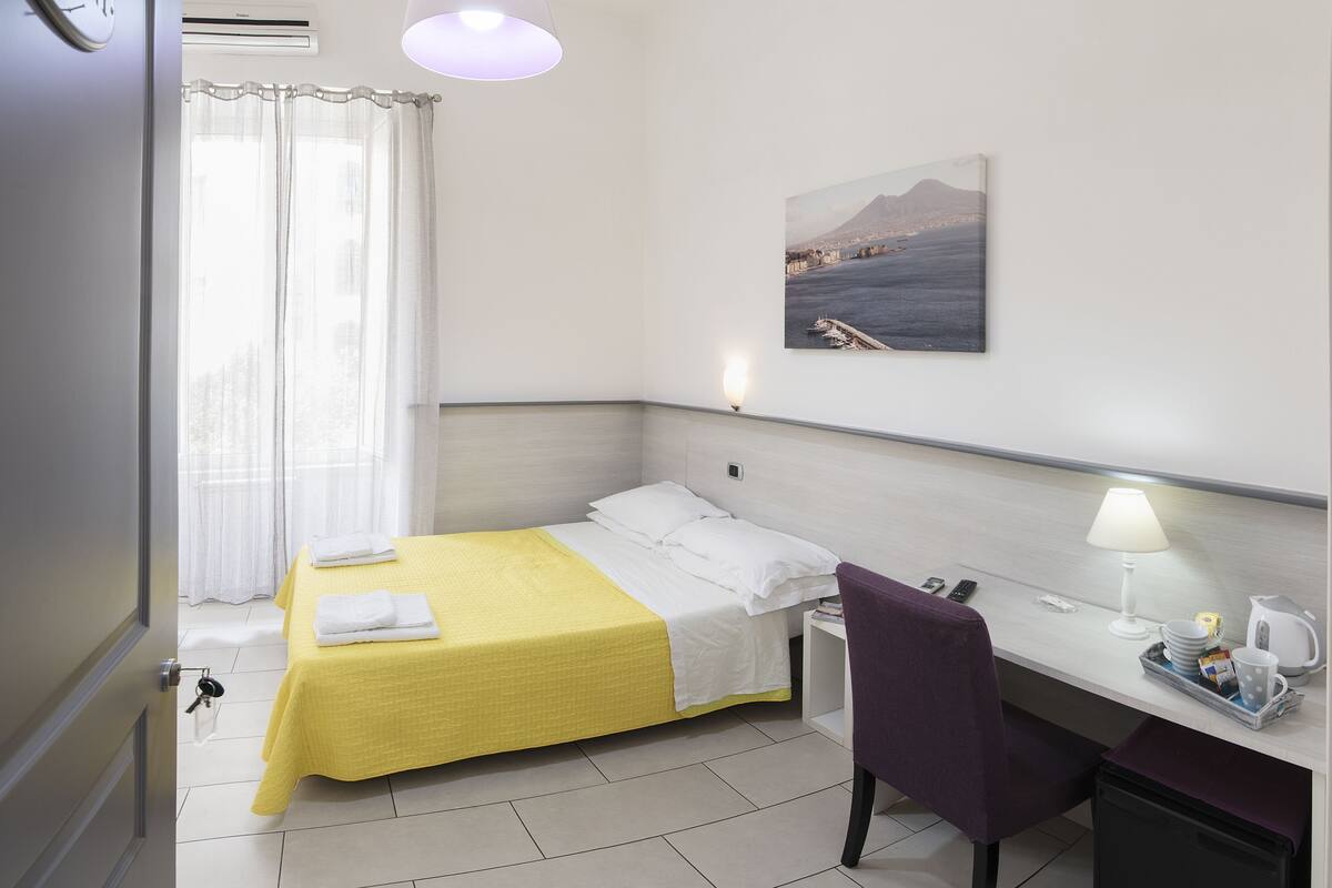 A hotel room with a bed, a desk, a chair, a window with curtains, a wall-mounted lamp, and a painting of a coastal scene.