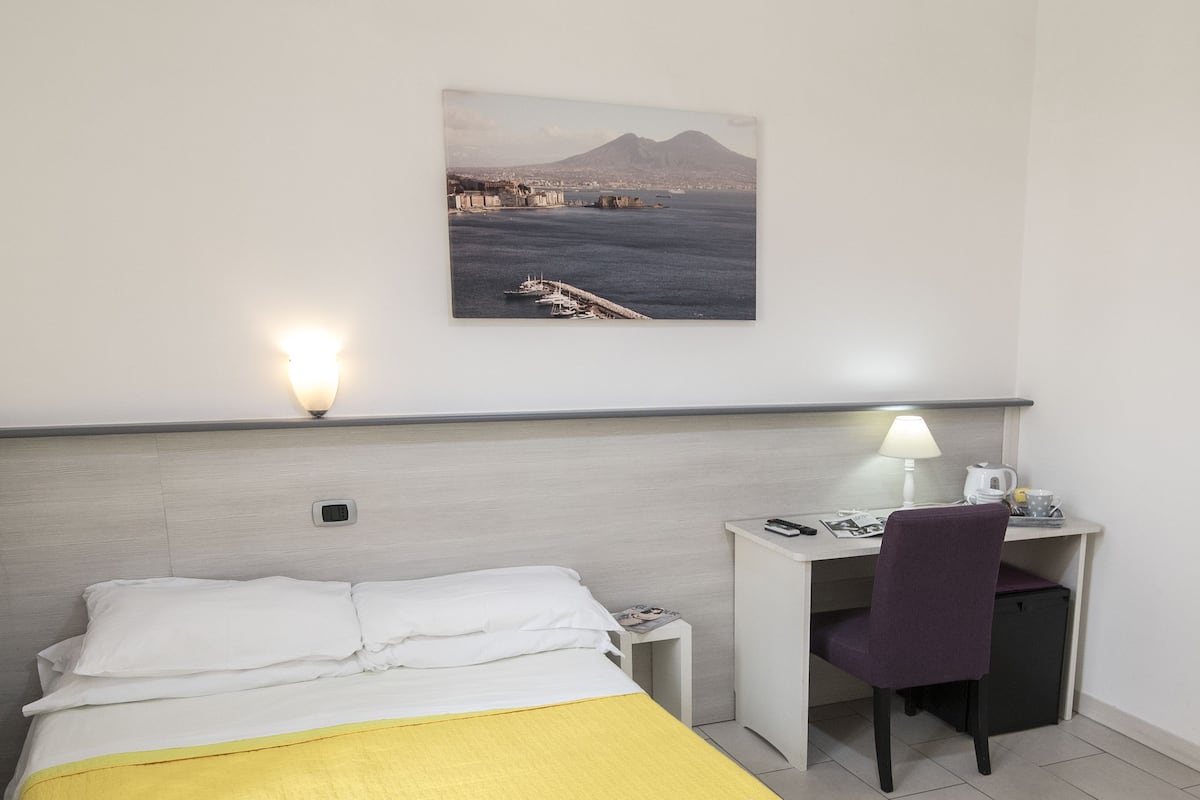 A hotel room with a bed, a desk, a chair, a lamp, and a painting on the wall.