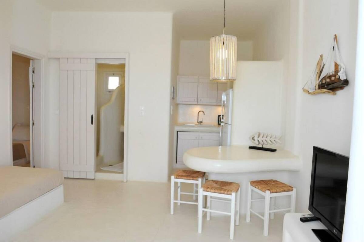 A compact room with a bed, a small dining area with two stools, a TV, and a kitchen area with cabinets and a hanging light fixture.