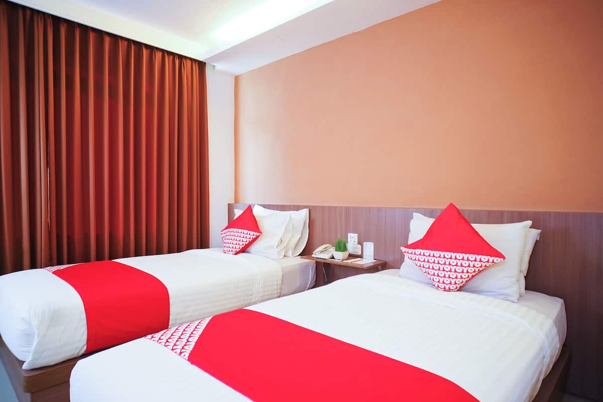 Duas camas com roupa de cama vermelha e branca, uma mesa de cabeceira com um telefone e uma planta, e uma janela com cortinas castanhas.