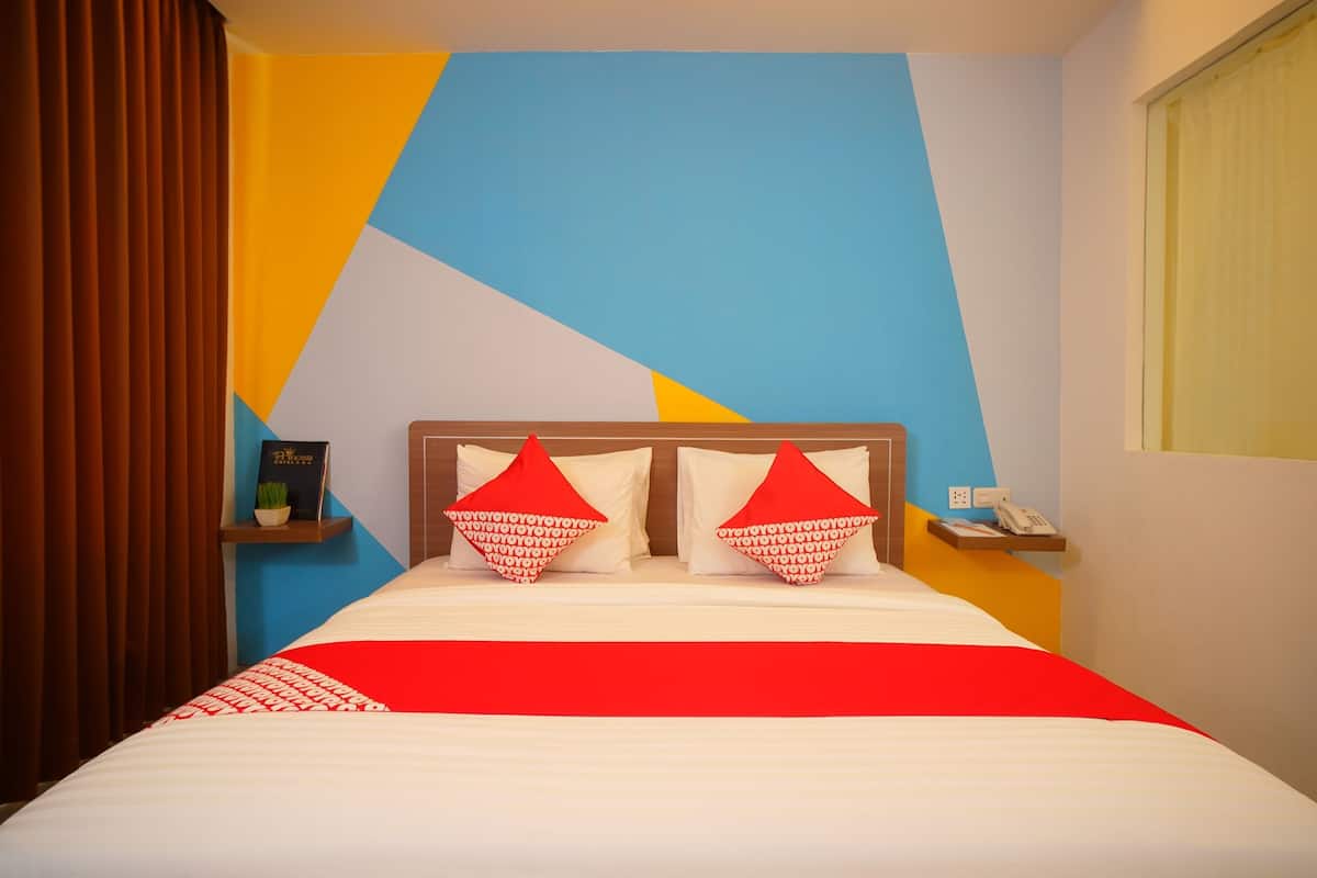 Quarto de hotel com cama, almofadas, mesa de cabeceira, relógio de parede e janela com cortinas.