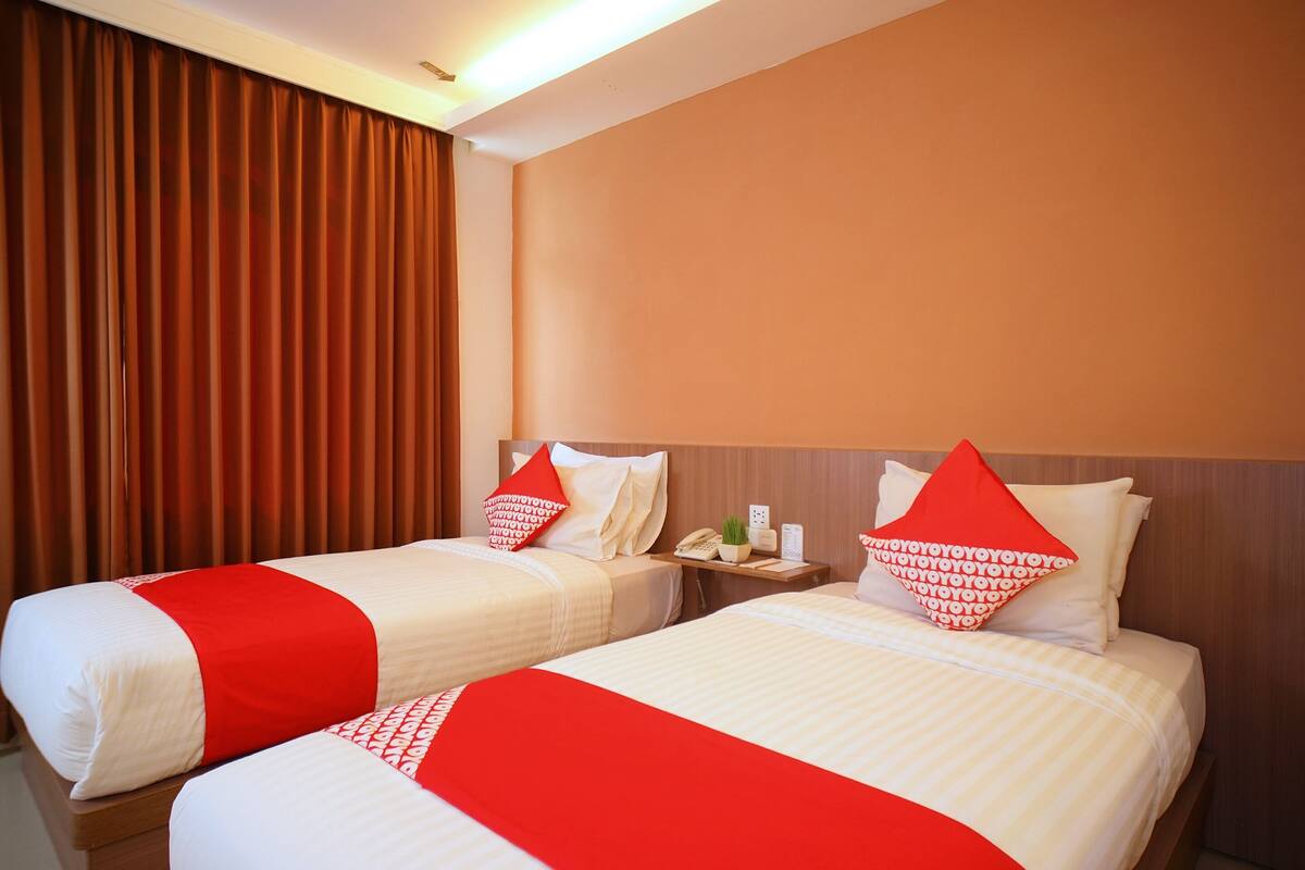 Duas camas com roupa de cama vermelha e branca, uma mesa de cabeceira com um telefone e uma planta, e uma janela com cortinas castanhas.