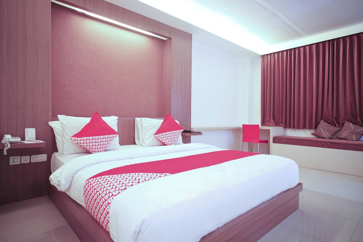 Quarto de hotel moderno com uma cama grande, mesa de cabeceira com telefone, escrivaninha com cadeira e janela com cortinas.