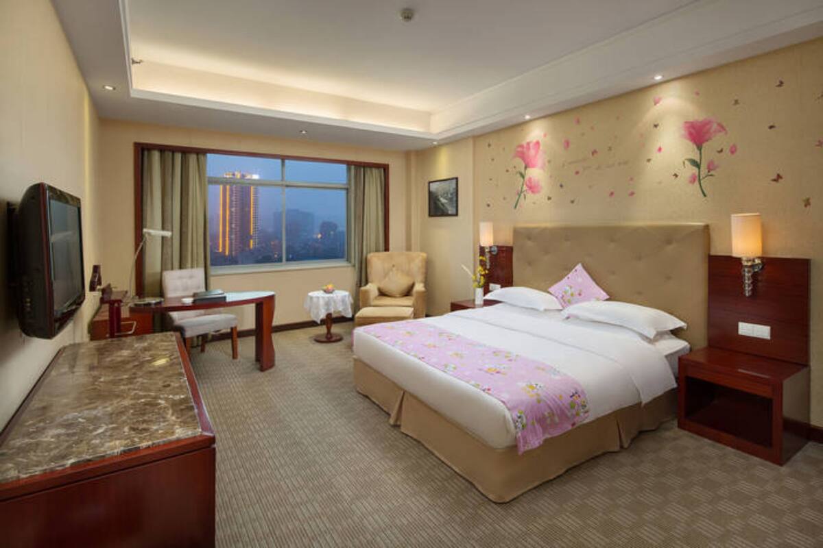 A hotel room with a large bed, a desk, a chair, a TV, and a window with a city view.