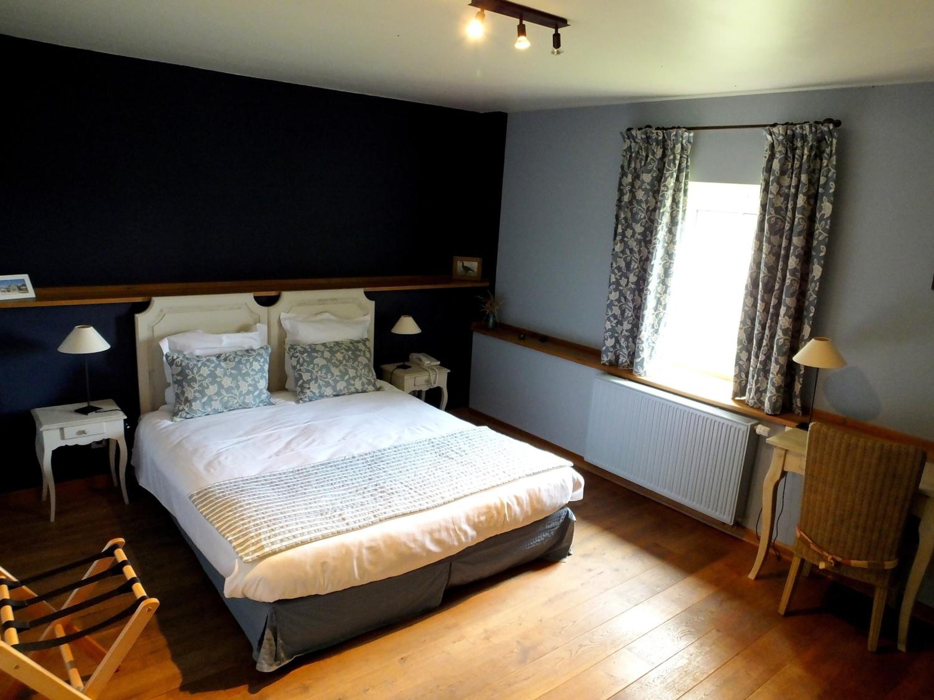 Une chambre à coucher avec un lit, des tables de chevet, une chaise, une fenêtre avec des rideaux et un radiateur.