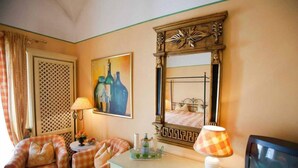 A hotel room with a checkered sofa, a small table with a lamp, a painting of bottles, a mirror with a decorative frame, and a bed with a metal frame. - Landgasthof Alte Bergmühle (Dreieich)