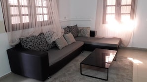 A living room with a dark-colored sofa, a glass coffee table, and sheer curtains on the windows. - Peniel Residence (Abidjan)