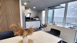 Un espace repas moderne avec une table blanche, deux vases contenant des plantes séchées et une vue sur la mer grâce à de grandes fenêtres.