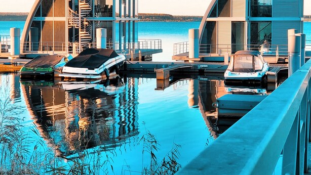 Une propriété moderne située au bord de l’eau, dotée de deux structures aux lignes courbes, d’un quai avec des bateaux et offrant une vue dégagée sur l’eau.
