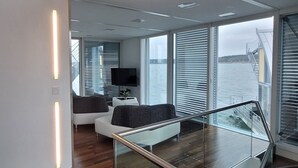 Un intérieur moderne avec un escalier en verre, un coin salon doté d’un téléviseur et de grandes fenêtres donnant sur l’eau.