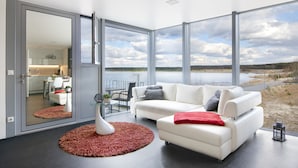 Un salon moderne avec un canapé blanc, un plaid rouge et une grande fenêtre donnant sur un lac.
