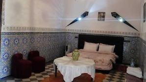 A room with a checkered floor, a bed with a pink bedspread, a white table with a vase, and a chandelier. - Riad Freija (Freyja)