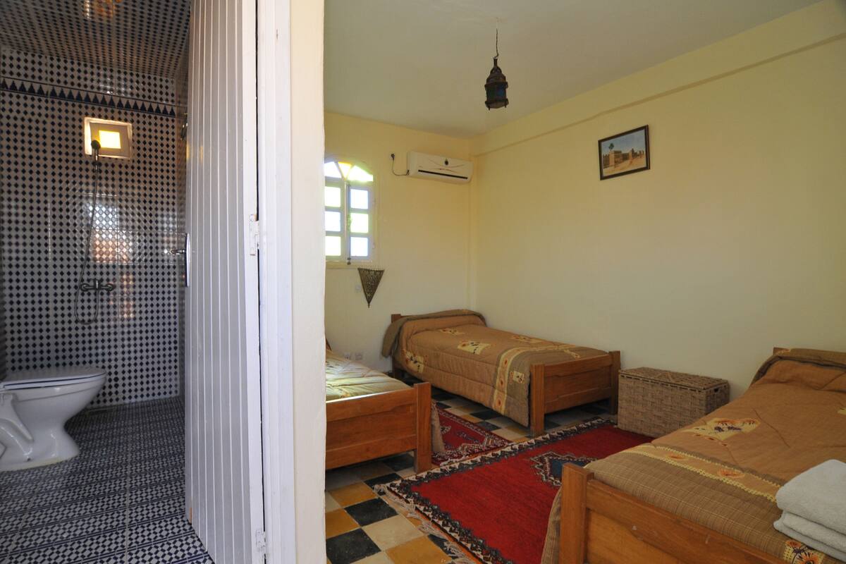 Una habitación con inodoro, dos camas, ventana, aire acondicionado y una lámpara colgante.