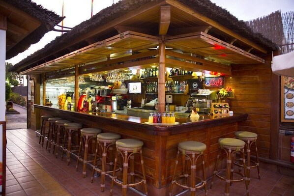 Un bar à l’extérieur en bois et au toit de chaume, avec un comptoir, des tabourets, ainsi que diverses bouteilles et verres.
