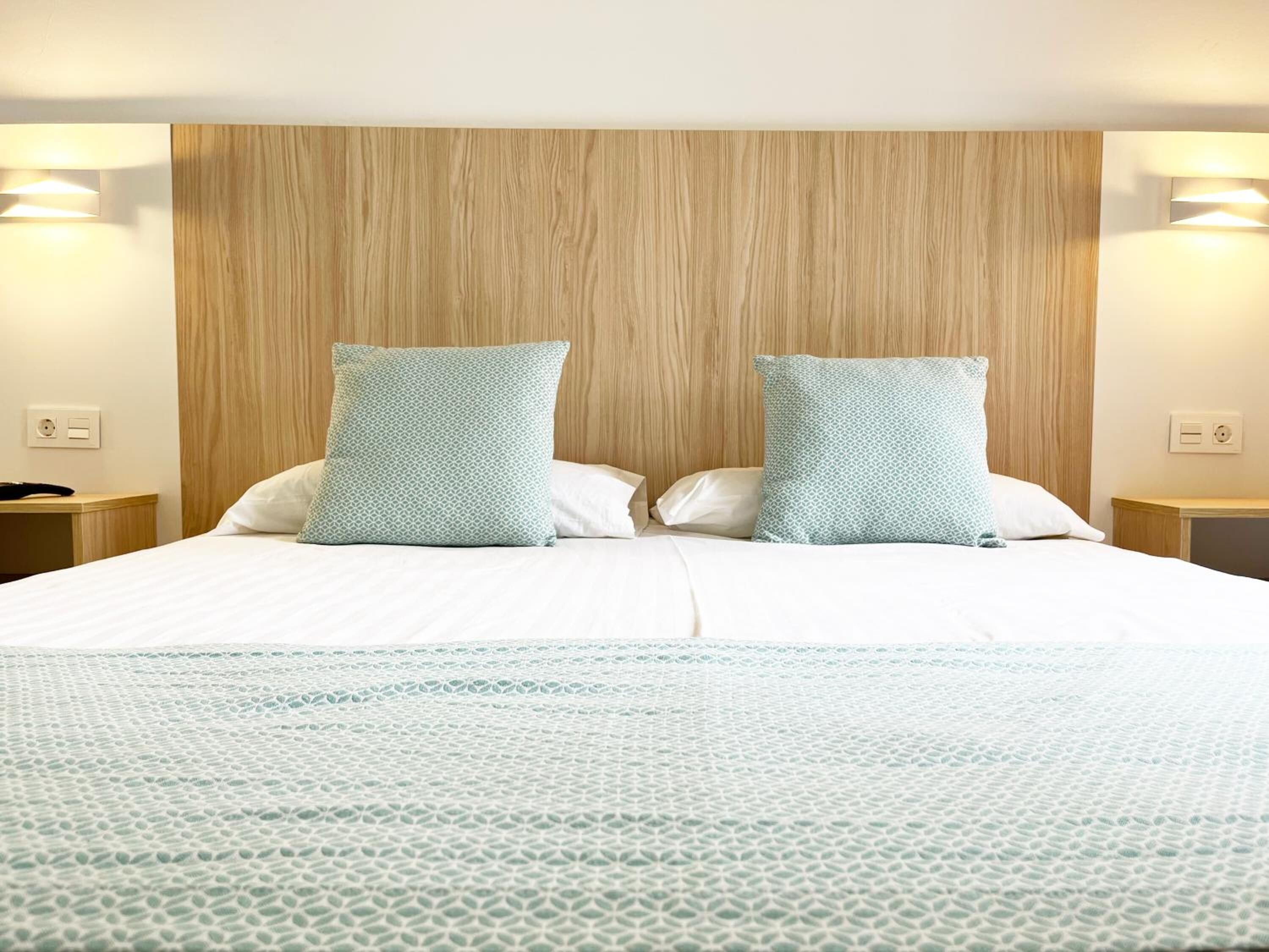 Ein Hotelzimmer mit einem Holzkopfteil, zwei Kissen mit geometrischem Muster, einer weißen Bettdecke und einem Nachttisch mit einer Lampe.