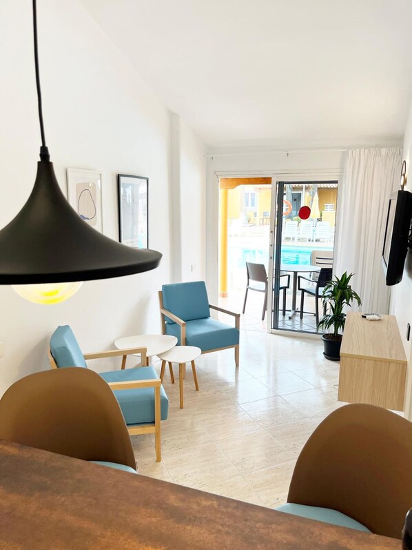 Un salon moderne comprenant un canapé, des fauteuils, une table basse et un coin repas avec une table et des chaises. Une grande fenêtre offre une vue sur une piscine et un espace de détente extérieur.