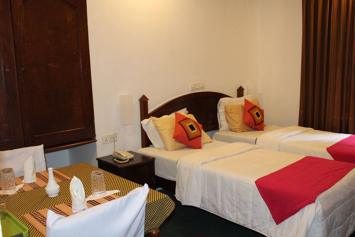 A hotel room with two single beds, a wooden headboard, a small bedside table with a phone, and a table with a placemat, glasses, and a vase.