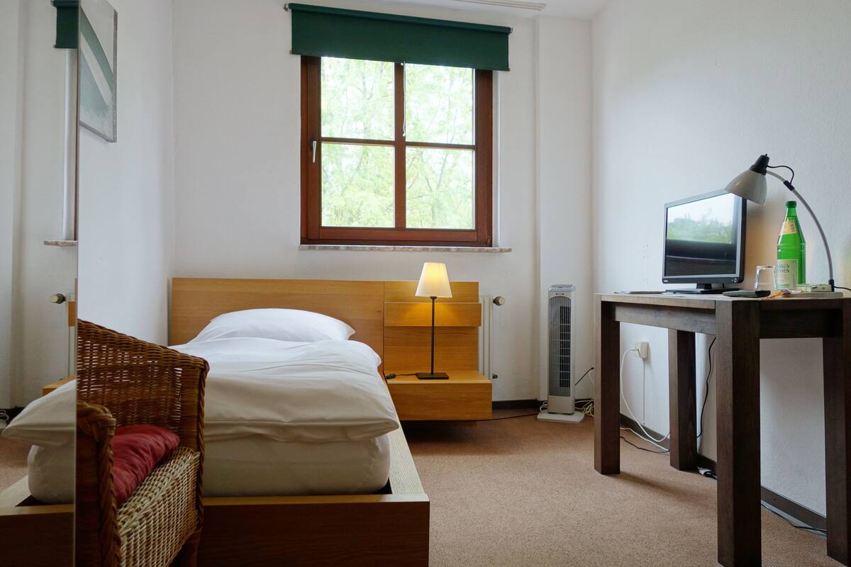 A neatly arranged hotel room with a bed, a bedside lamp, a desk with a computer and a bottle, and a window with a view of trees.