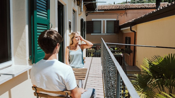 A balcony with a wooden table and chairs, a metal railing, and a view of a building with a tiled roof and green shutters.