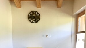 Een kleine kamer met een houten plafond, een wandklok, een tafel met twee stoelen, een deur en een raam met gordijn.