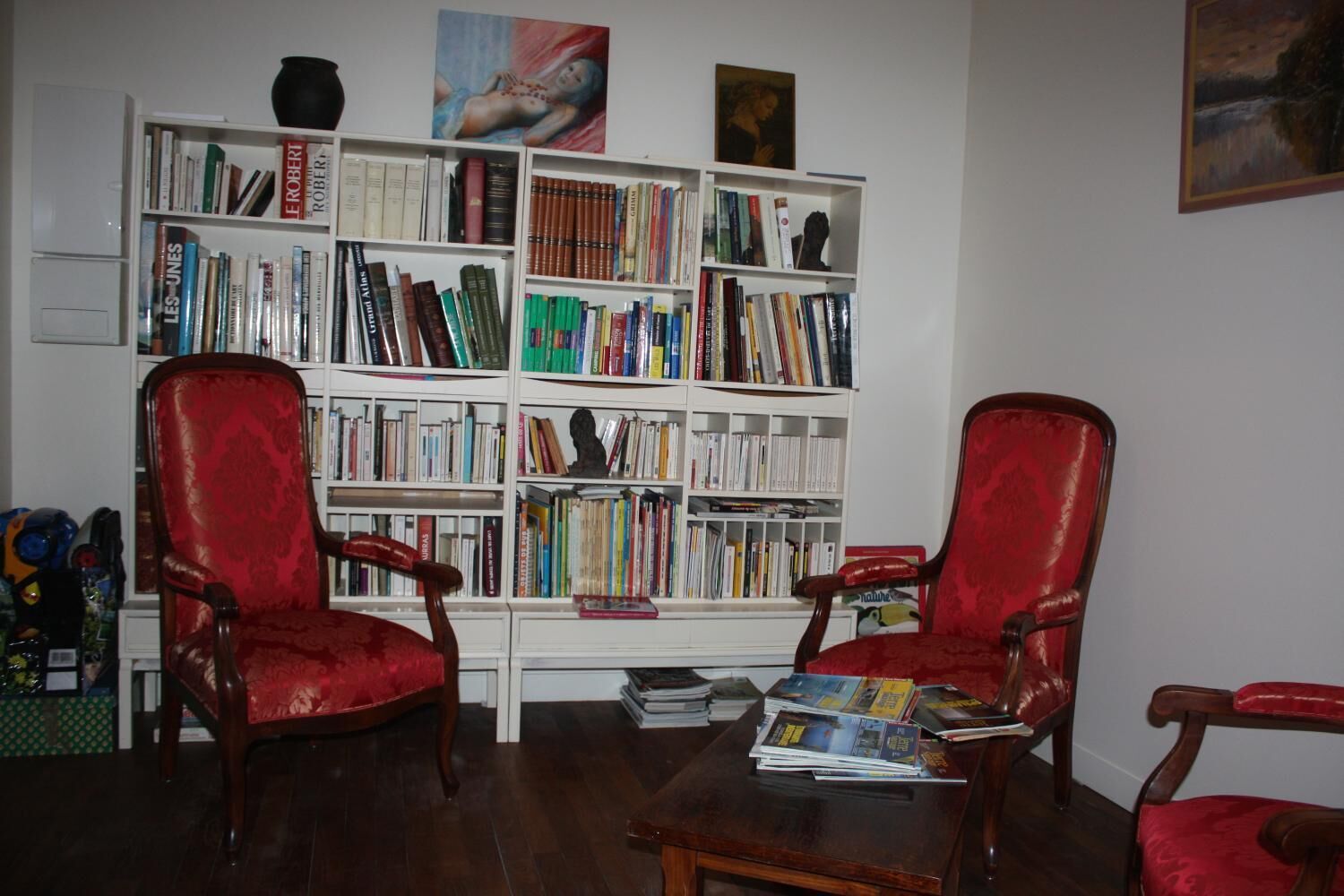 Una habitación con una amplia estantería blanca llena de libros, dos sillones tapizados en rojo y una mesa de centro de madera.